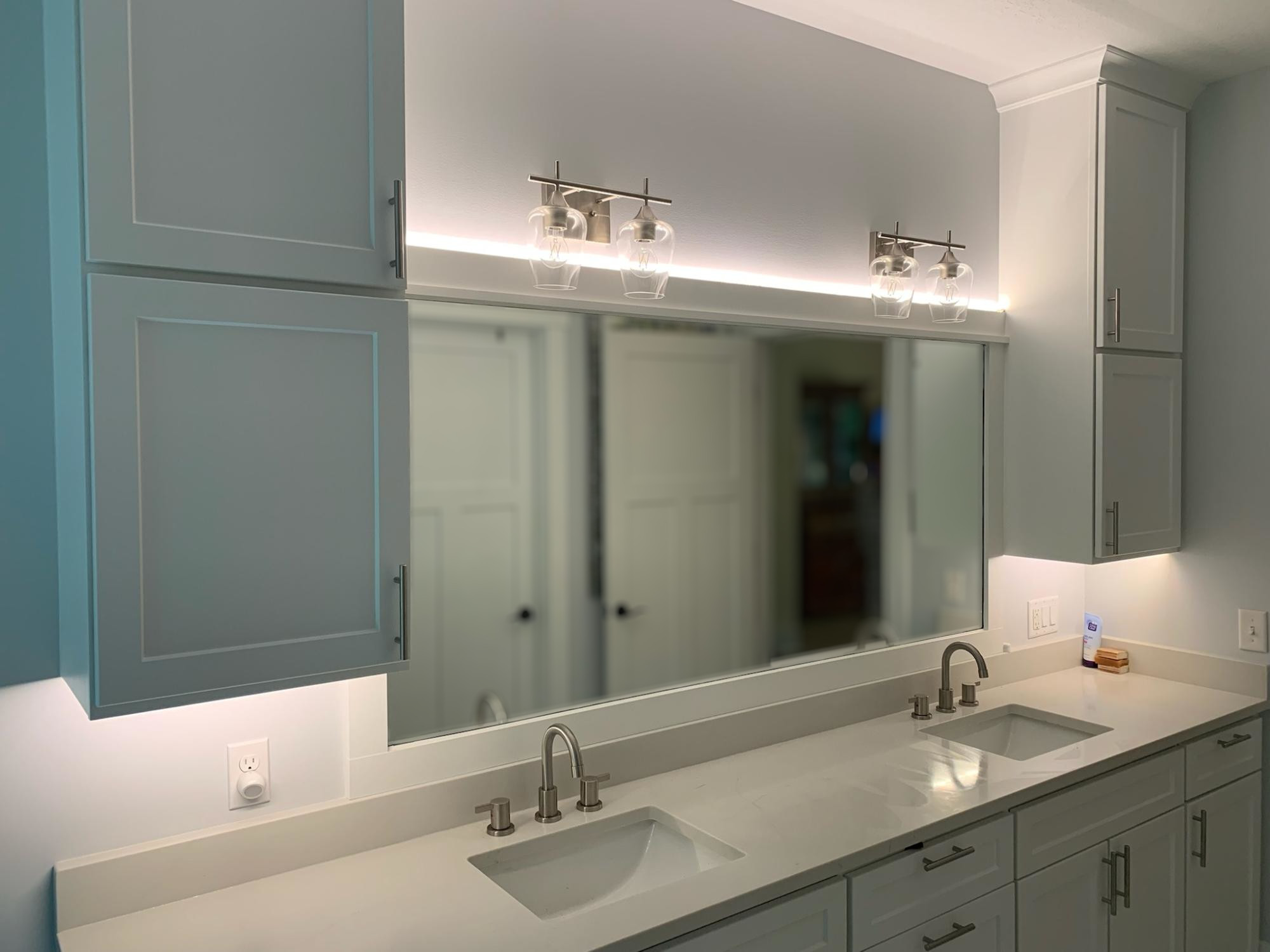 A clean, bright bathroom with nice lighting.  There is a man in the mirror's reflection, standing oddly in the distance, hidden behind a false blur.